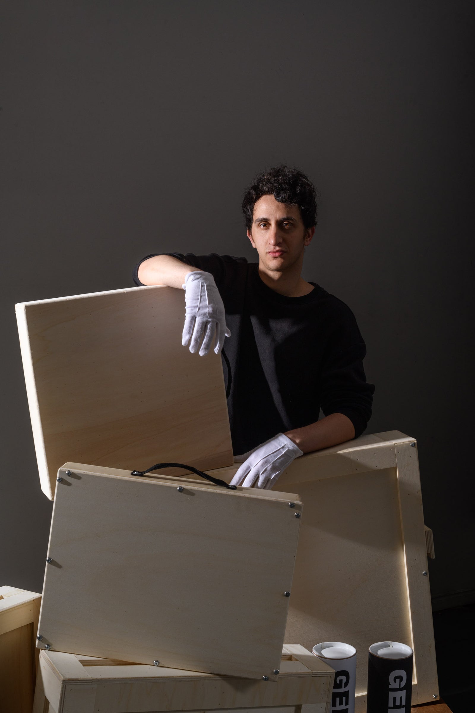 A man in white gloves stands behind several wooden art crates, holding one open while looking directly at the camera. Three GEMS-branded tubes are stacked beside him on the floor.