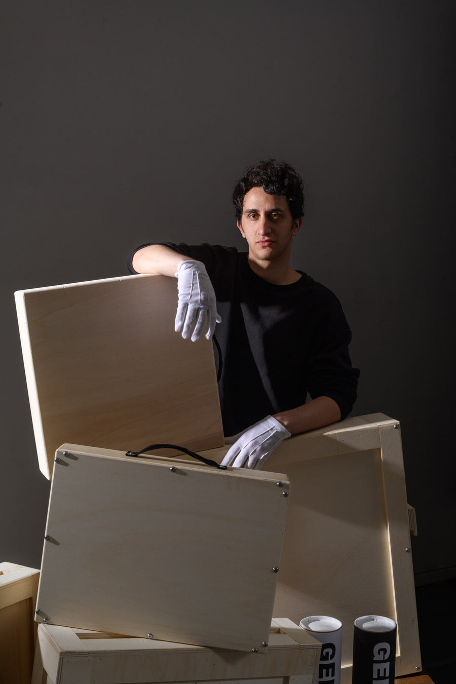 A man in white gloves stands behind several wooden art crates, holding one open while looking directly at the camera. Three GEMS-branded tubes are stacked beside him on the floor.