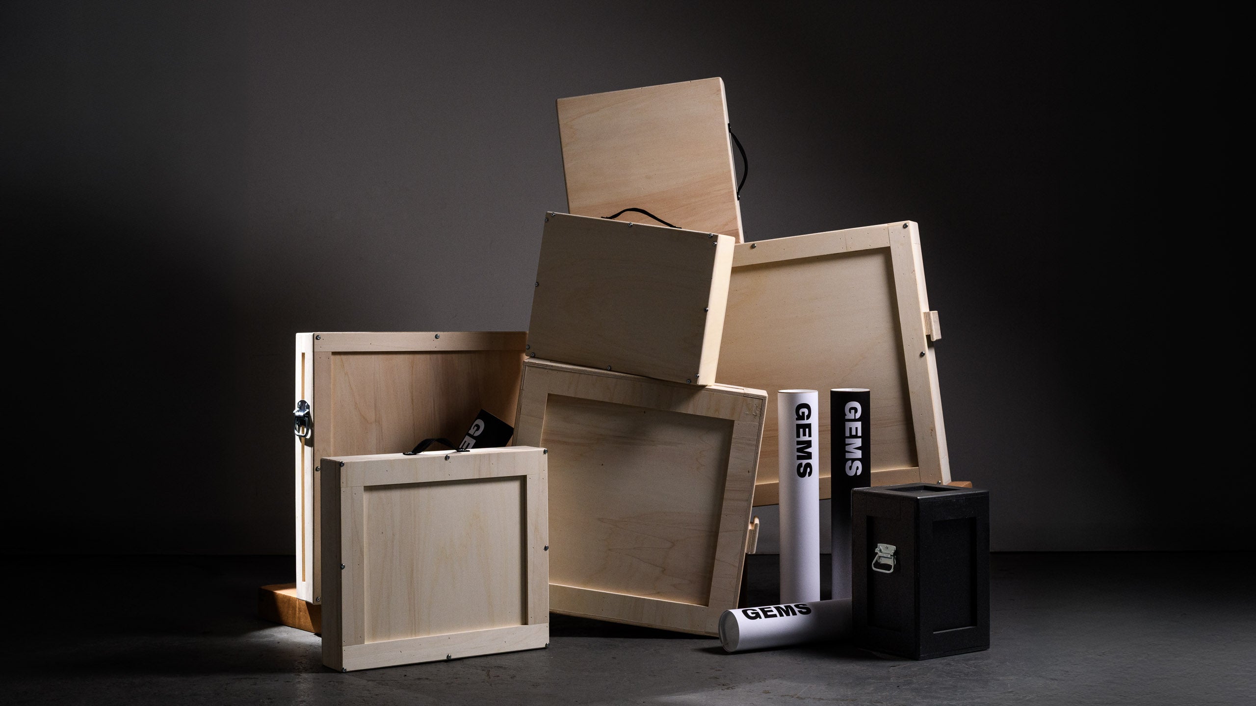 Group of stacked wooden and black crates with GEMS-branded poster tubes arranged in the foreground, dramatically lit against a dark backdrop to highlight premium art shipping solutions.
