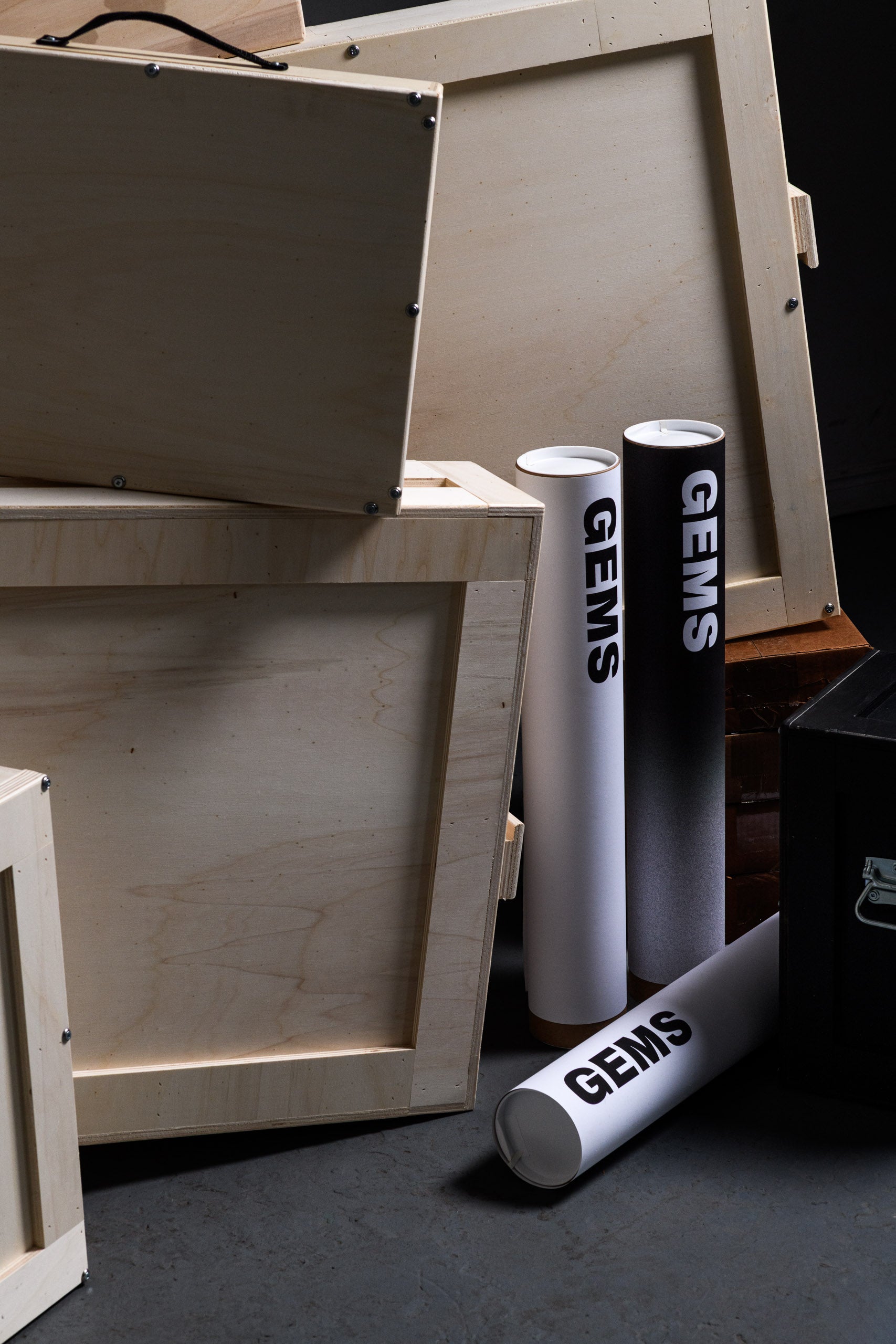 Close-up of three GEMS-branded poster tubes in white and gradient black, standing upright among open wooden art shipping crates, emphasizing professional packaging and branding.

