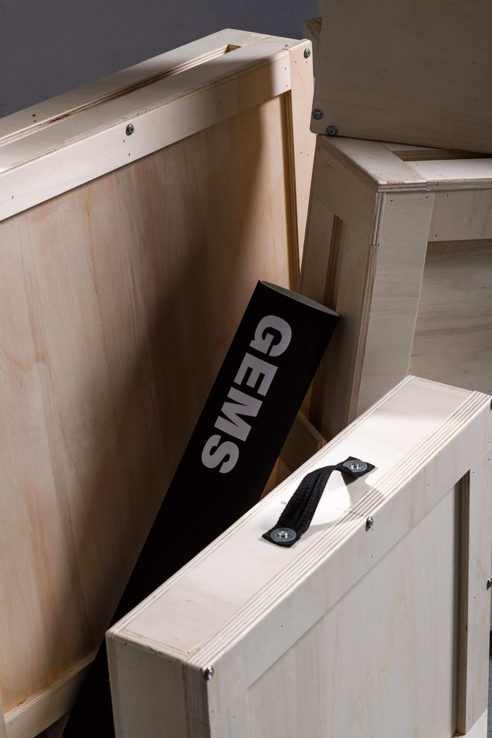A black GEMS poster tube partially visible between stacked wooden crates, highlighting the minimal, elegant branding and custom-built wooden packaging.

