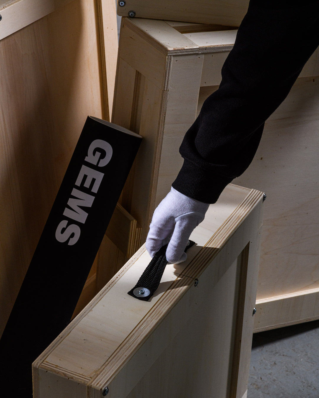 Gloved hand lifting a bespoke wooden crate with a durable black handle, conveying the safe handling and transport of collectible artworks.