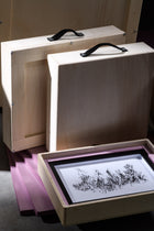 Two neatly stacked wooden crates with soft black handles, next to an open crate holding a framed monochrome drawing, all sitting on purple foam pads that cushion and protect art during shipping.