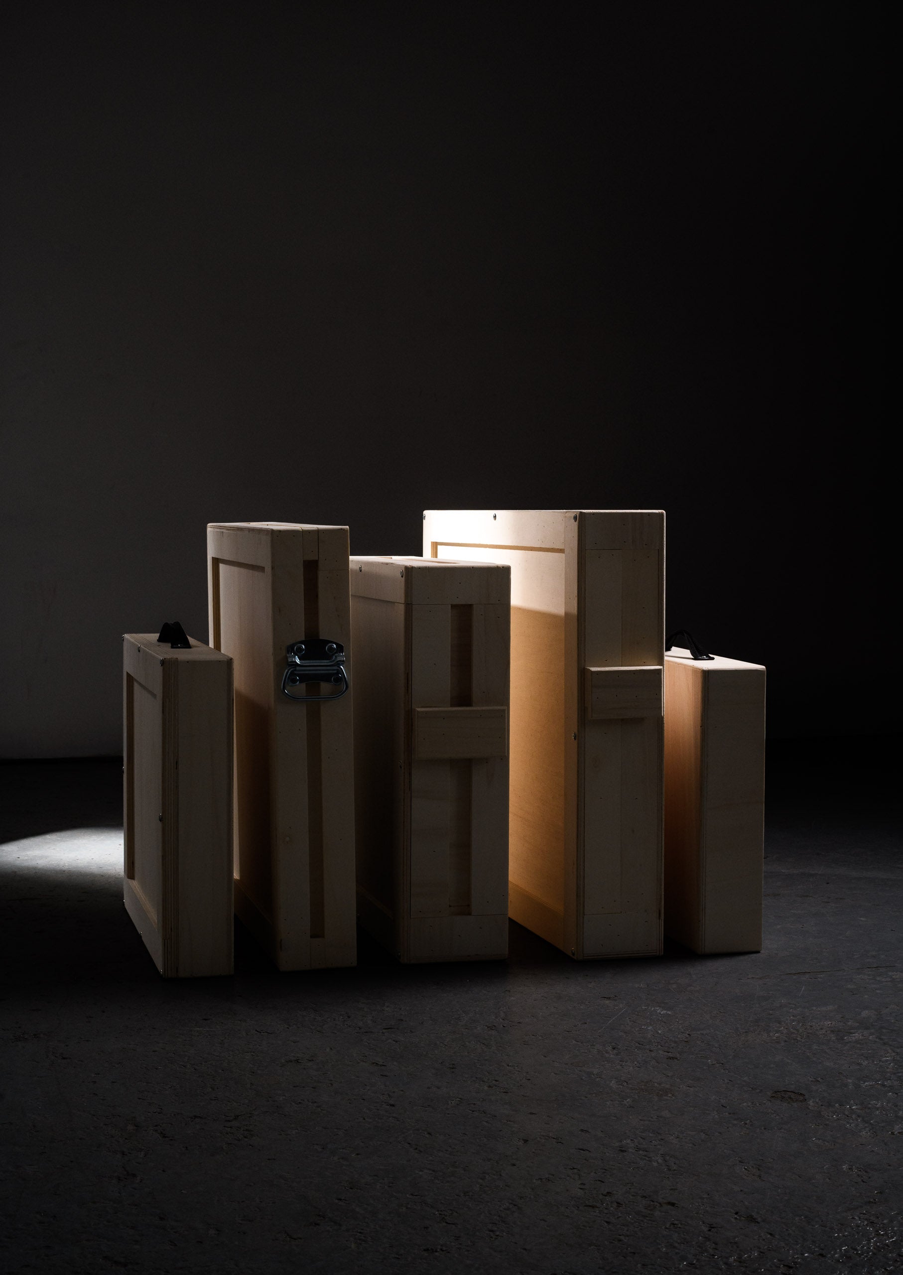A collection of premium wooden art crates arranged with a sculptural presence in a dark studio, illuminated by natural side light to reveal their protective structure.