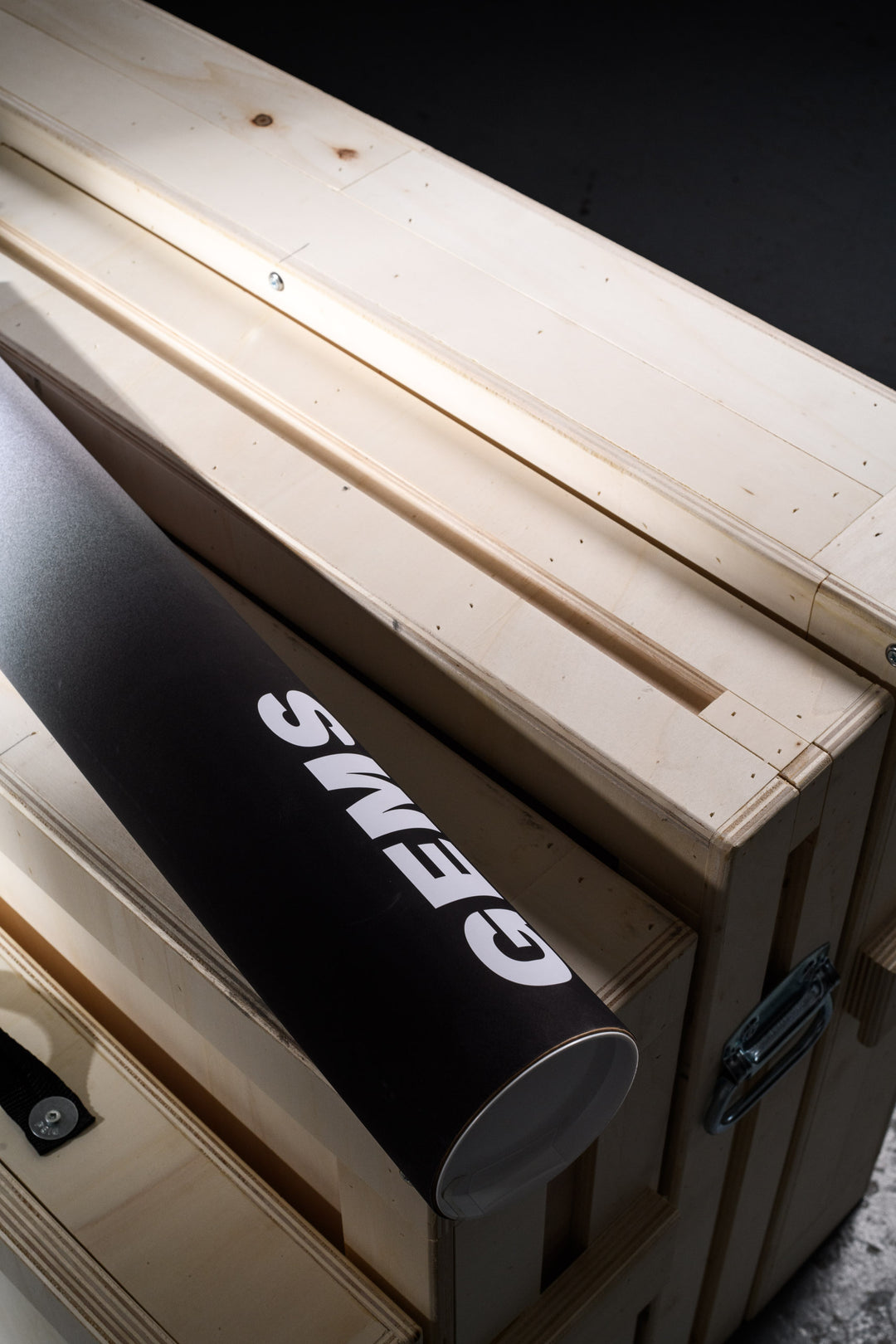 A black GEMS-branded print tube resting atop a wooden shipping crate, symbolizing the fusion of sleek presentation and professional art packaging.
