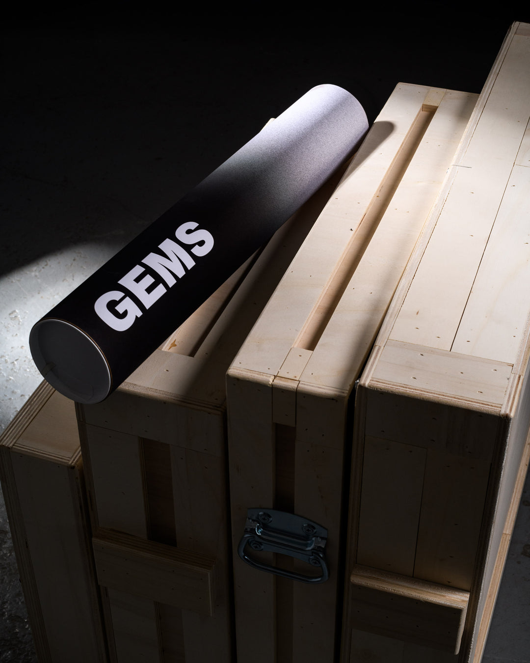 Top-down view of a branded GEMS print tube laid across multiple stacked wooden crates, emphasizing the clean design and premium handling of art logistics.
