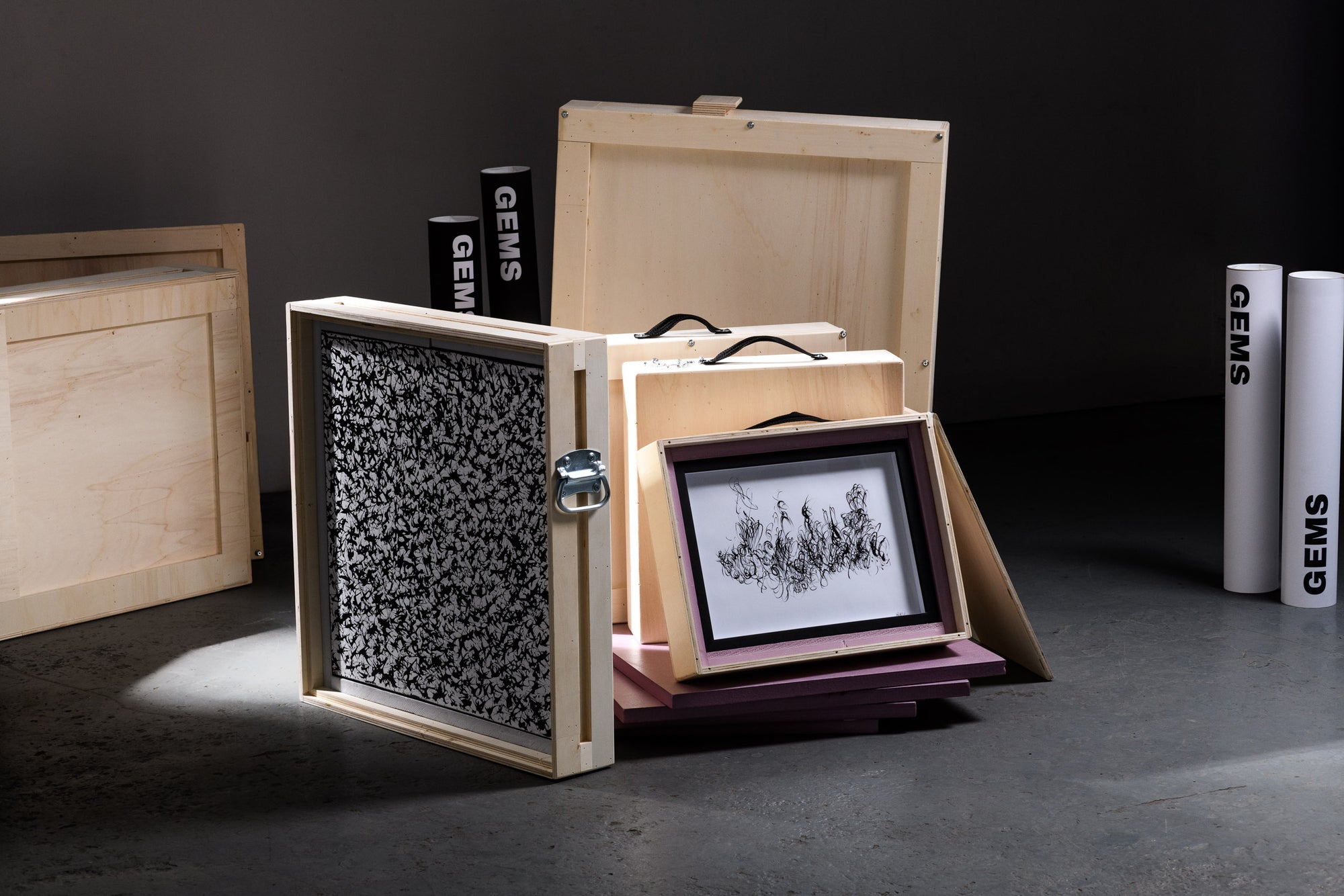 An elegant arrangement of open art crates revealing framed original works, surrounded by GEMS-branded tubes, representing the safe transport and curated delivery of collectible art.