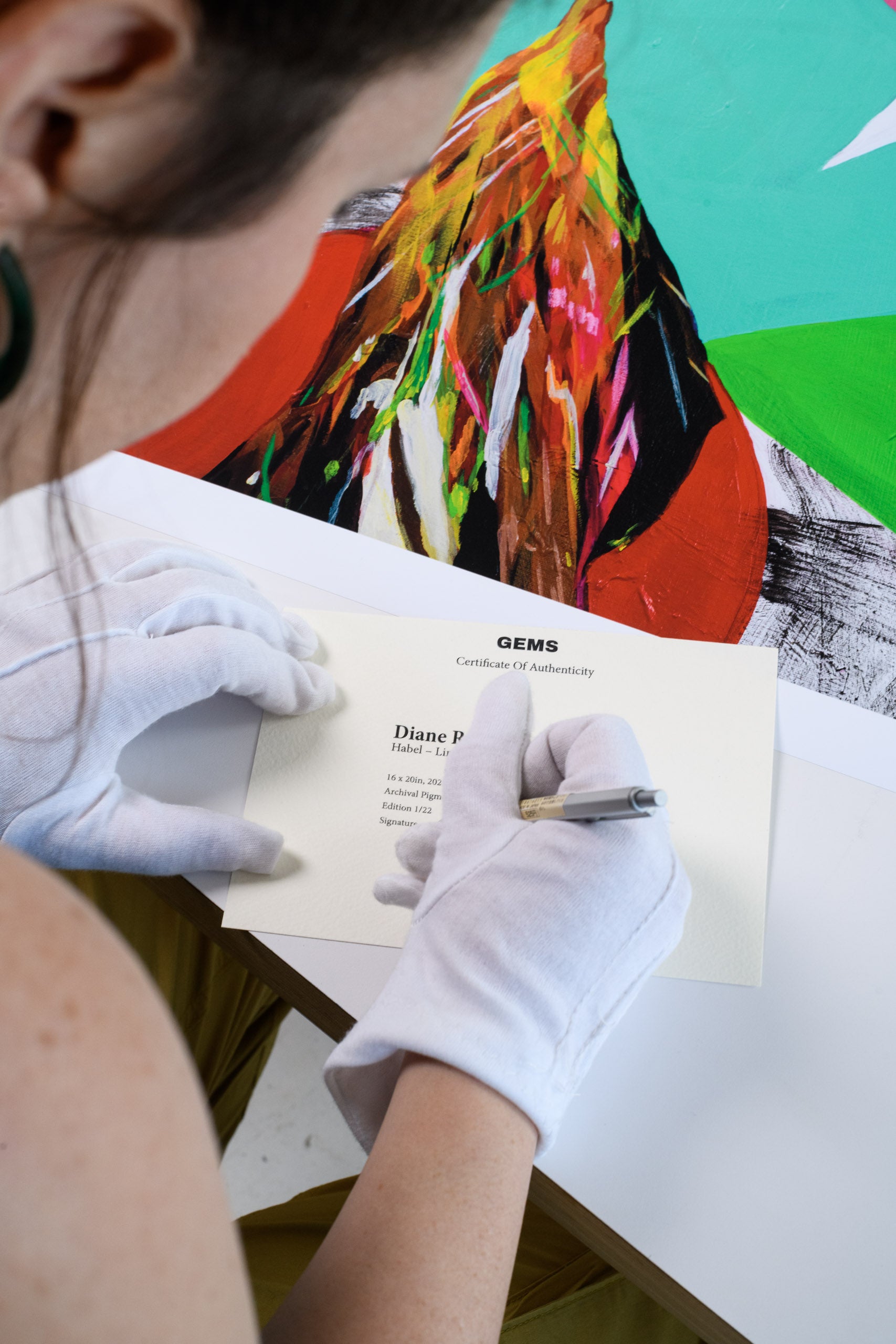 A woman wearing white gloves signs a GEMS certificate of authenticity next to a vibrant limited edition print featuring bold abstract colors.