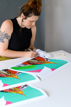  Artist carefully signs limited edition prints laid out on a white table, wearing white gloves to handle the artwork with care.