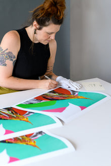  Artist carefully signs limited edition prints laid out on a white table, wearing white gloves to handle the artwork with care.