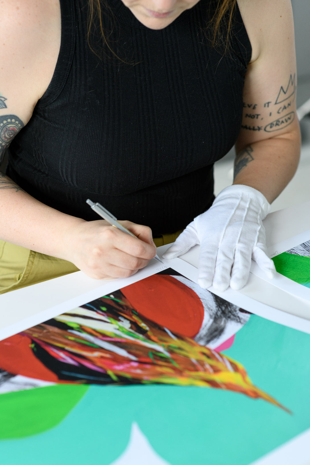 Close-up of a gloved hand signing the corner of a colorful print, marking the edition number with precision and care.