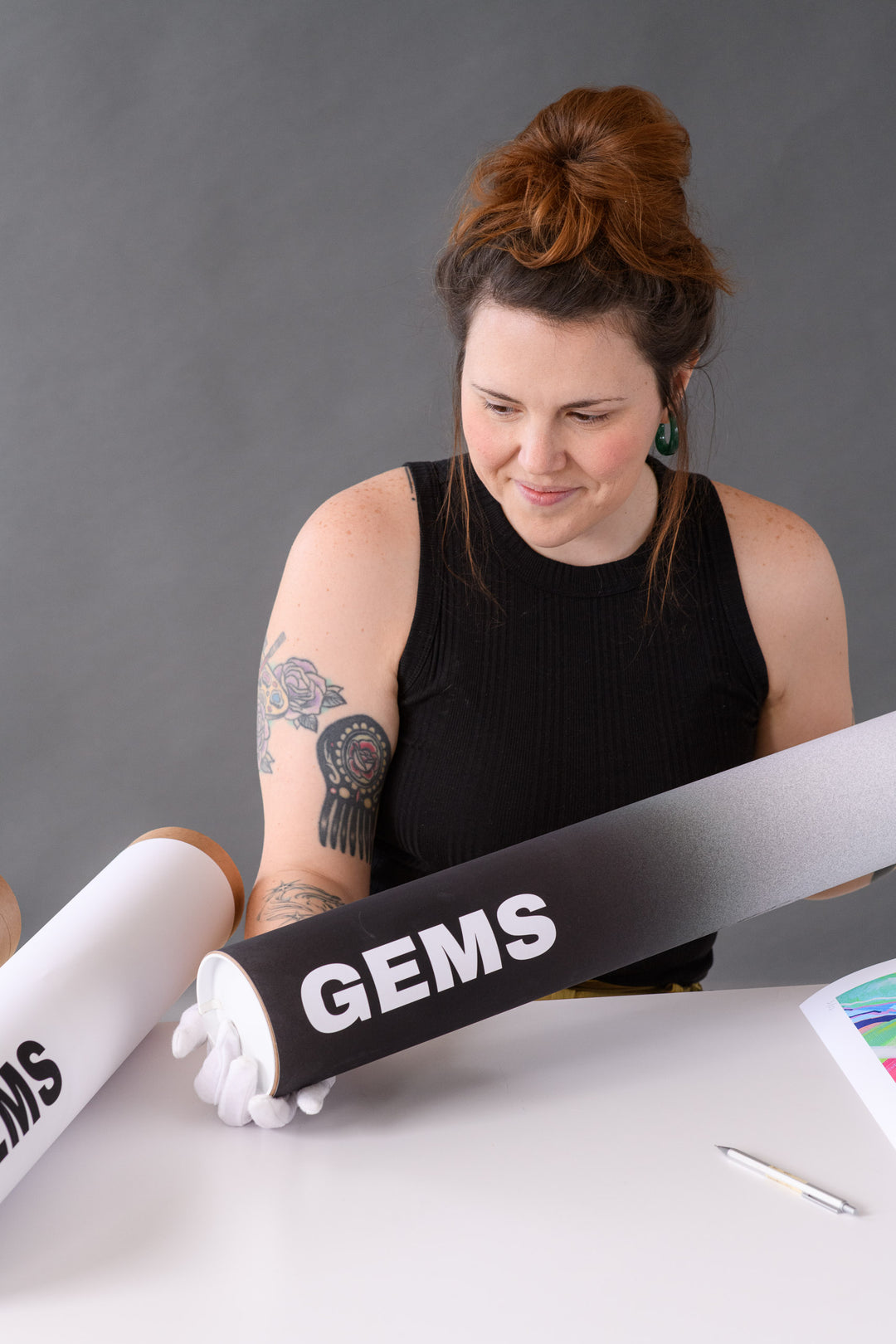 A smiling woman holds a branded GEMS art tube with white gloves, ready to package a signed limited edition print.
