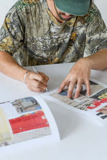 A man in a camouflage shirt and cap signs the corner of a limited edition art print with precise attention to detail.