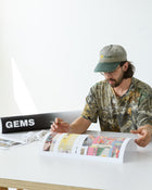 Artist inspects a series of colorful collage prints at a clean workstation, with a branded GEMS art tube and white gloves nearby.