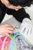 Person wearing gloves and using a magnifying glass on a colorful artwork