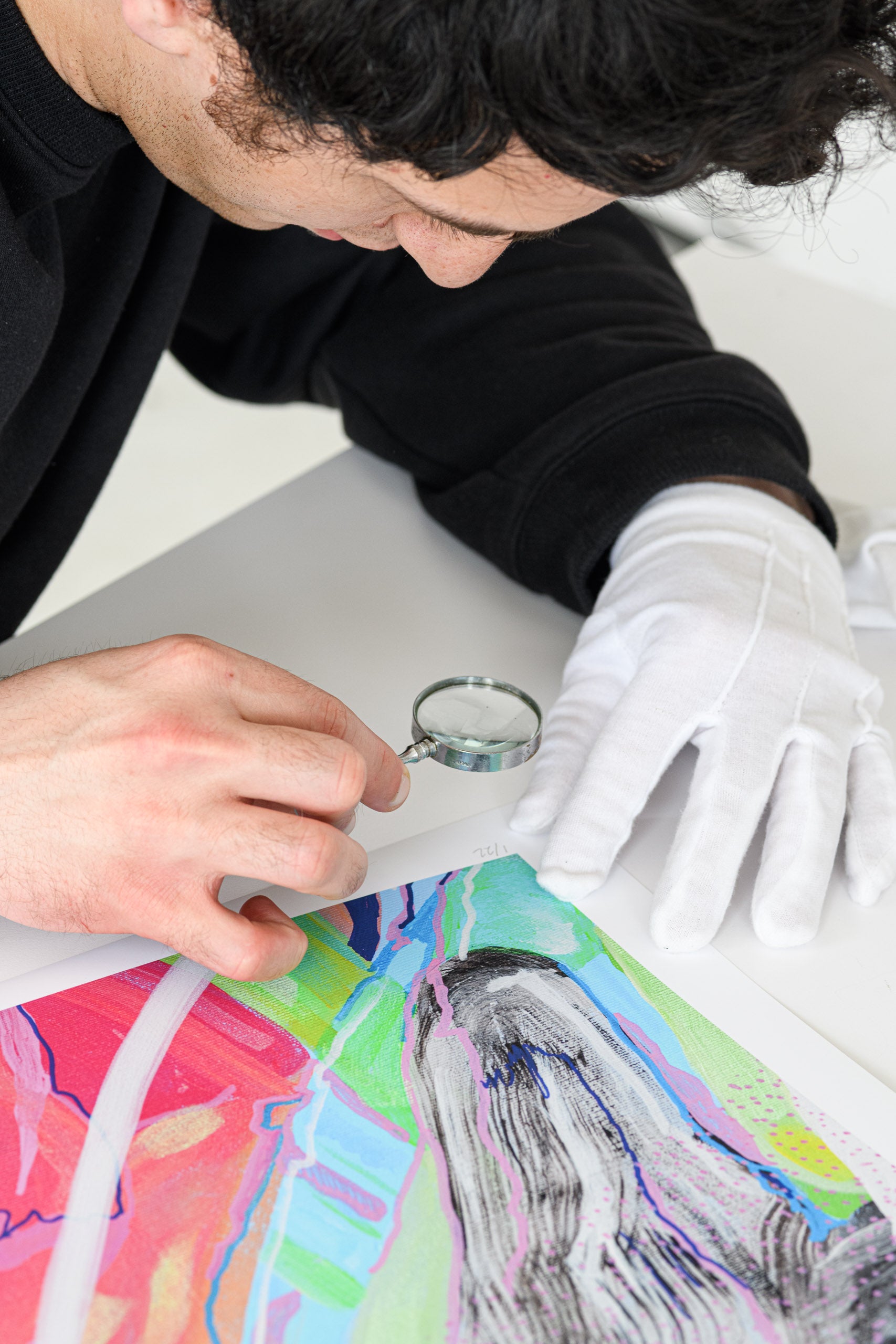 Person wearing gloves and using a magnifying glass on a colorful artwork