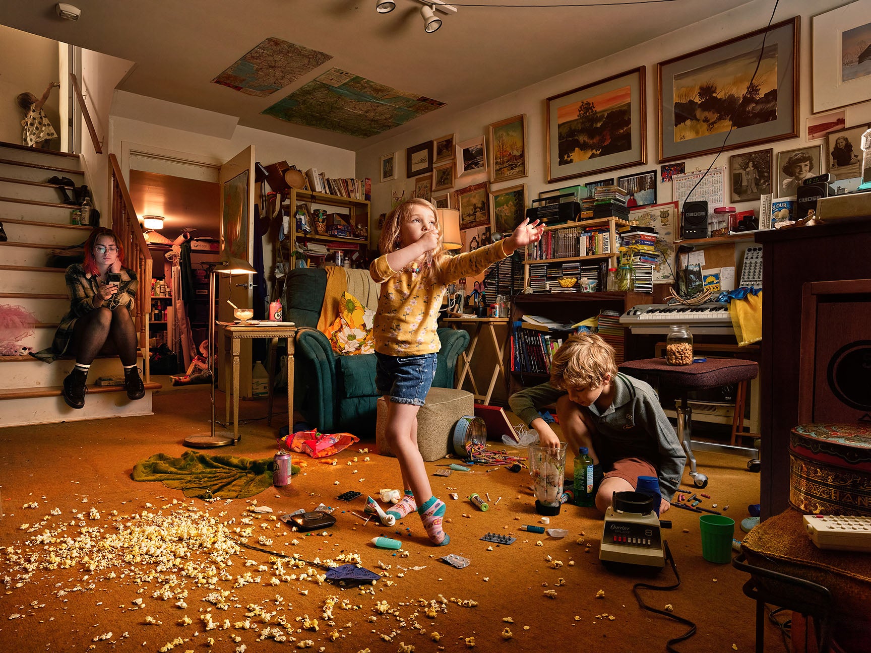 Three people in a cluttered room with books, furniture, and scattered items.